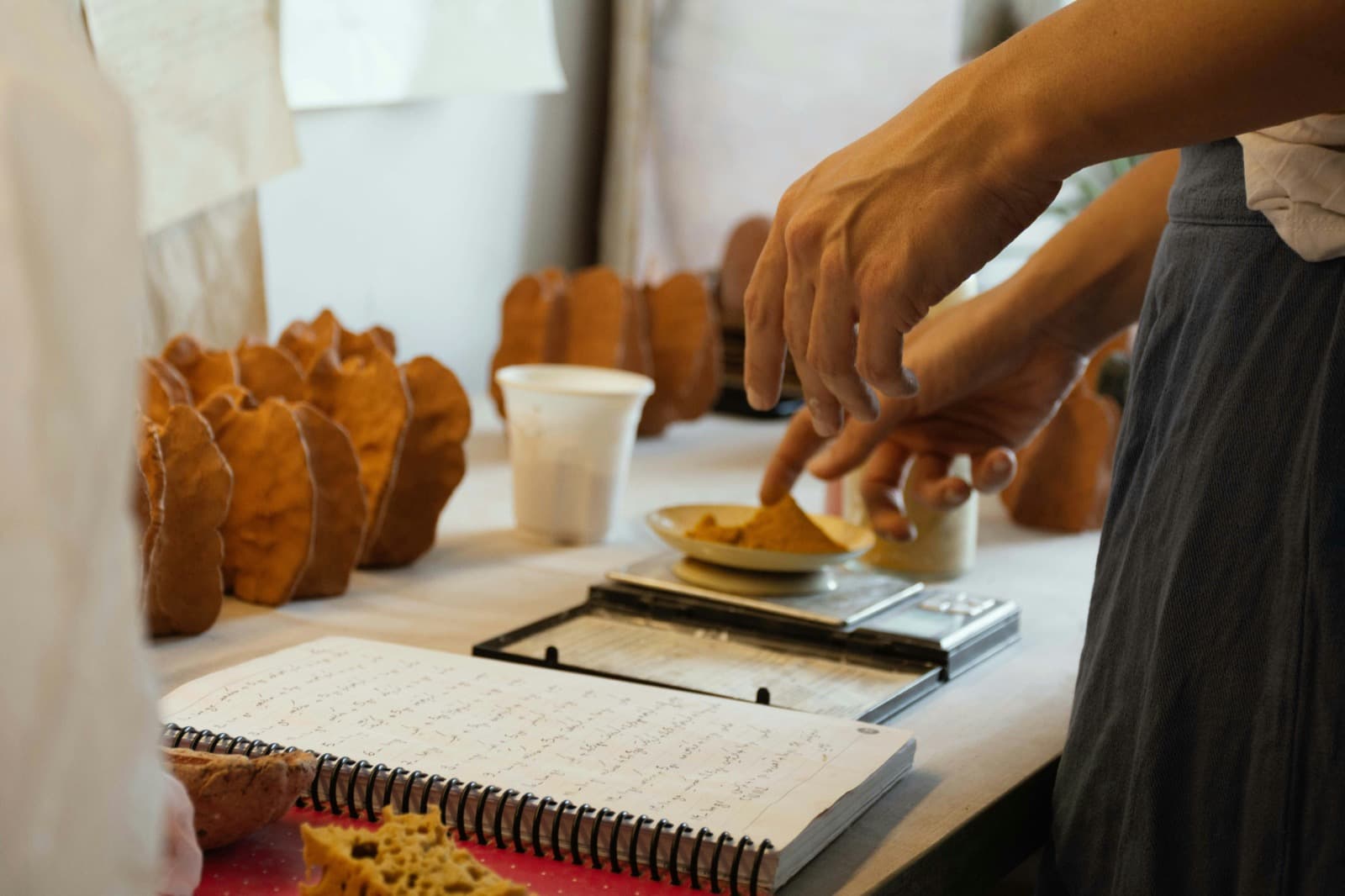 Manos pesando ingredientes en una balanza de cocina, junto a un cuaderno con anotaciones